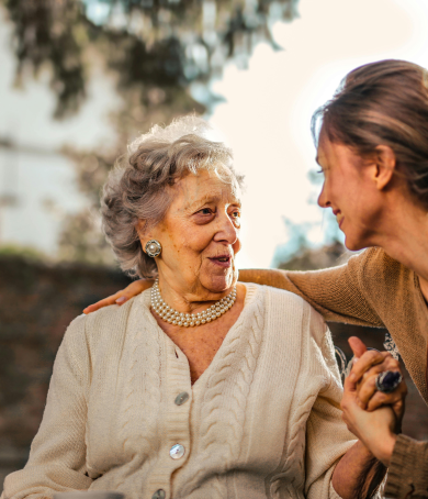 Elderly woman smiling and talking with a middle-aged woman outdoors, with a graphic icon symbolizing 'Holistic Support' and text overlay. Both women display joy and trust, emphasizing a nurturing, supportive conversation.